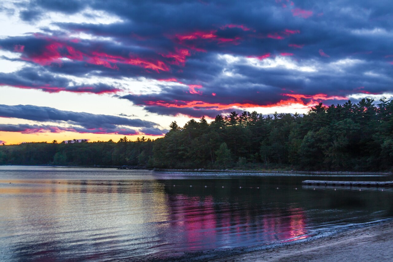 A vivid sunset over Sebago Lake, with a forested shoreline reflected in calm water